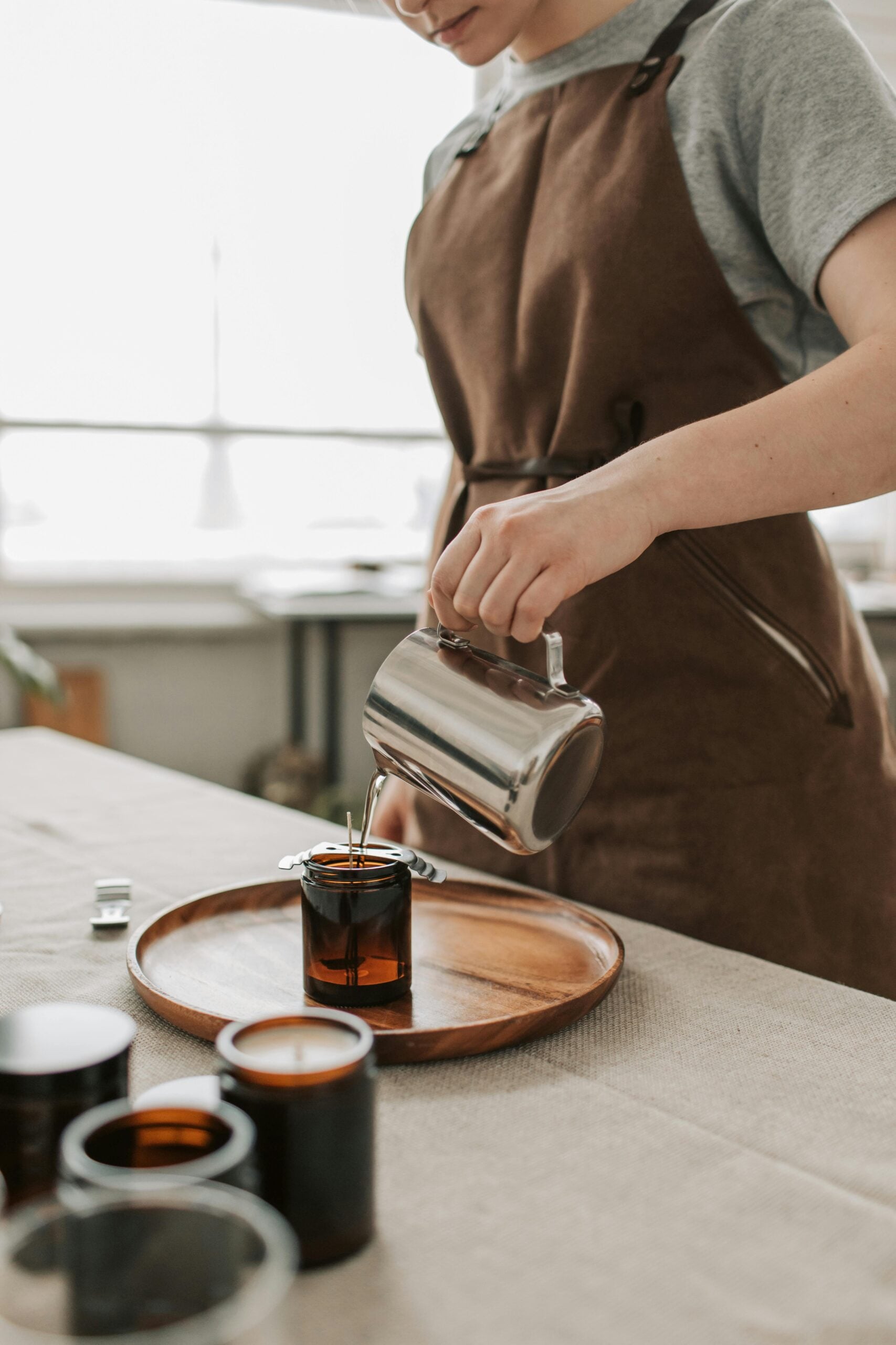 Craftsman in apron pours hot wax into a brown glass jar for candle making indoors.