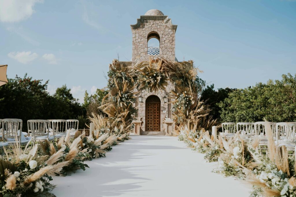pexels-photo-14716281-14716281-1 Beautiful outdoor wedding aisle with floral decorations leading to a stone building under a clear sky.